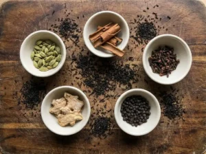 Whole spices for chai tea drink mix arranged in small bowls — green cardamom pods, Ceylon cinnamon sticks, whole cloves, dried ginger, and black peppercorns.