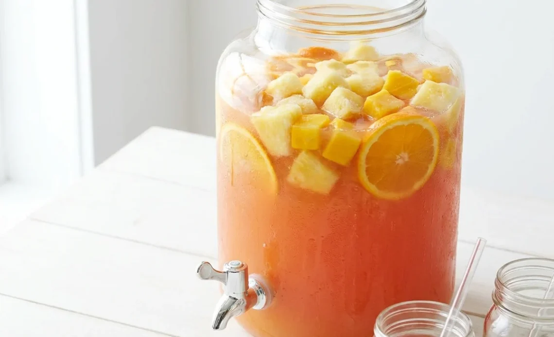 Jungle juice recipe in a glass dispenser with frozen tropical fruit and citrus slices