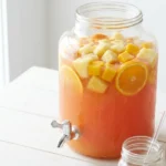 Jungle juice recipe in a glass dispenser with frozen tropical fruit and citrus slices