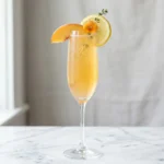 peach bellini mocktail in champagne flute with golden peach color peach slice and edible flower garnish on white marble