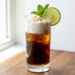 Coconut lime dirty soda in a tall Collins glass with a creamy coconut cream layer on top, lime wheel and toasted coconut garnish