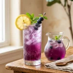 Blueberry lemonade in a tall glass with ice, lemon wheel and blueberries beside a pitcher