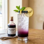 Blueberry simple syrup in a glass bottle beside a glass of iced blueberry lemonade with lemon and mint