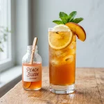 Peach syrup in a glass bottle beside a glass of iced peach tea with fresh peach slices and mint